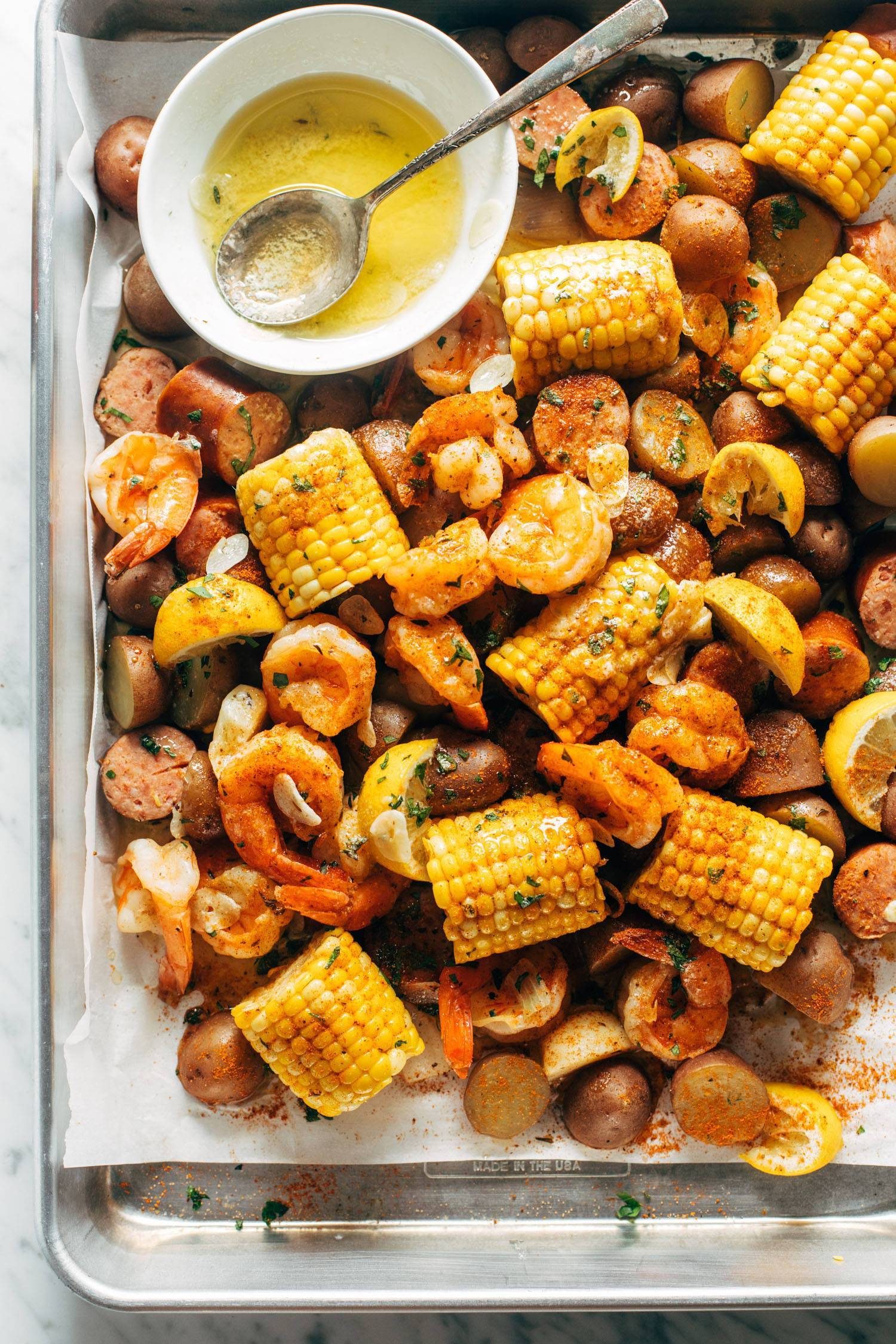 Shrimp boil on a sheet pan with garlic butter. The garlic butter is in a bowl with a spoon and there's corn, lemon wedges, shrimp, and kielbasa on the sheet pan with potatoes.